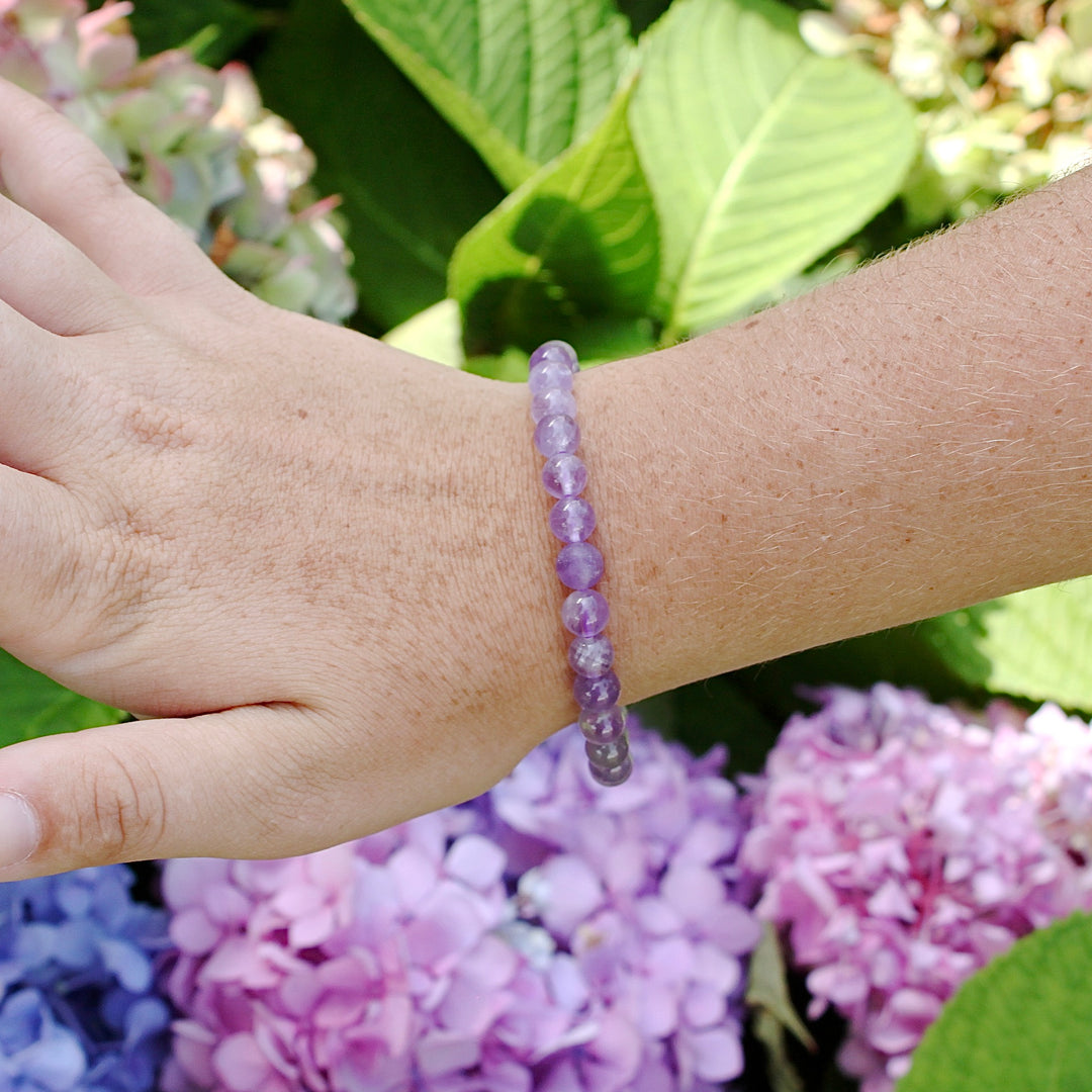 Lavender Amethyst 6mm Beaded Gemstone Bracelet on a wrist with flowers.